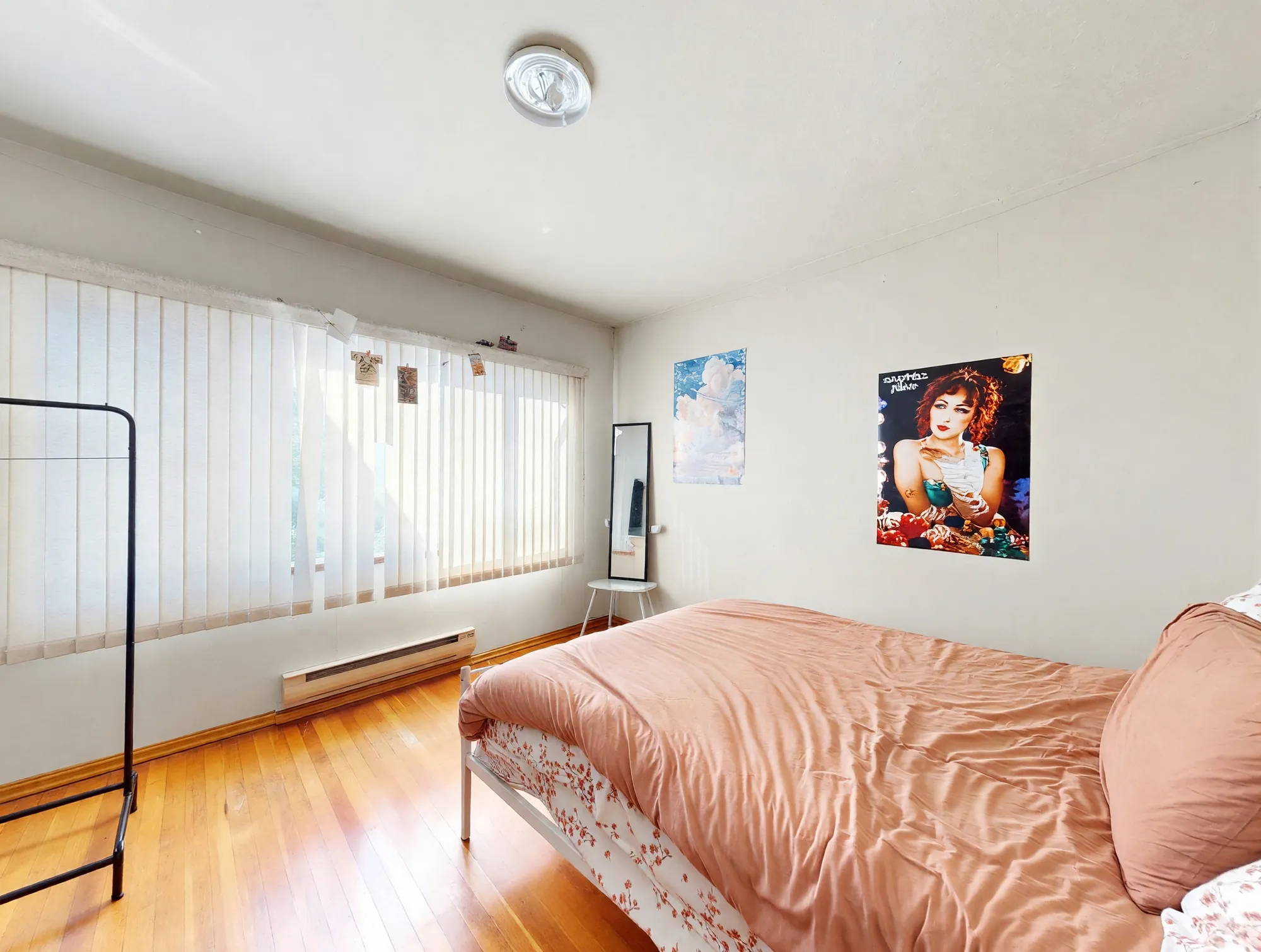 A bedroom with hardwood floor and a wide window behind vertical blinds